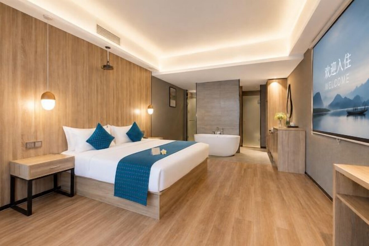 Une chambre d’hôtel moderne dotée d’un grand lit, de tables de chevet, d’une baignoire intégrée et d’une grande fenêtre offrant une vue dégagée.