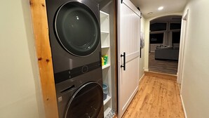 A compact laundry room with a front-loading washer and dryer, a white door, and a hallway leading to a living area. - Spacious, private and peaceful 1st floor Apartment on Fly Creek  (Fly Creek)