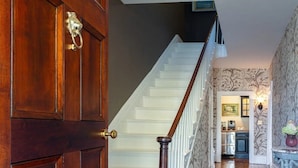 Una puerta de madera con manija de bronce, una escalera con barandillas blancas y una pared con papel tapiz estampado.