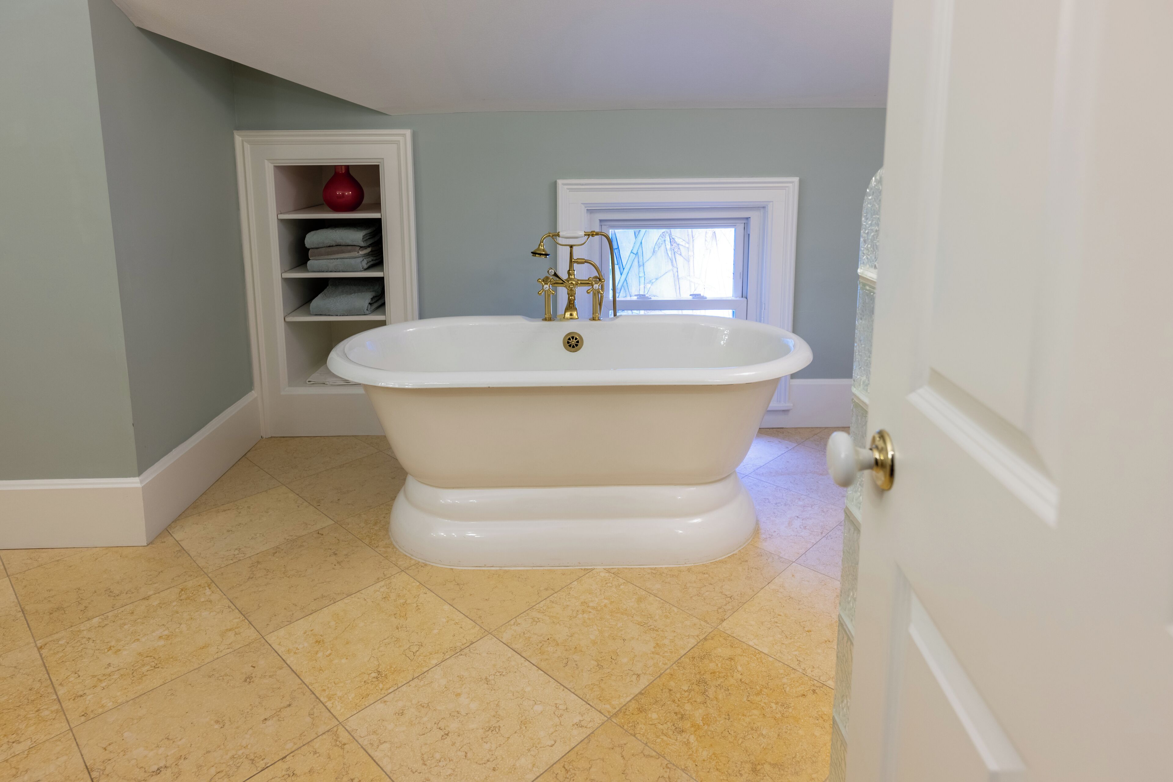 A bathroom with a freestanding bathtub, a window with a view, and a shelf with towels.