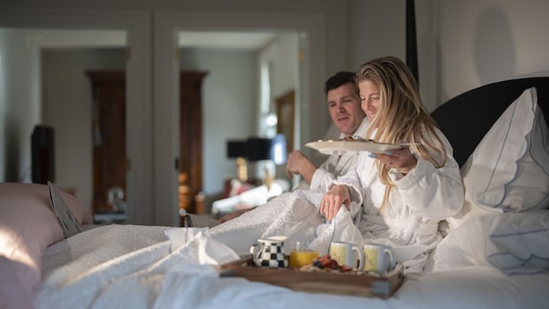 Una pareja en una habitación de hotel compartiendo una bandeja de desayuno con café, fruta y bollería.