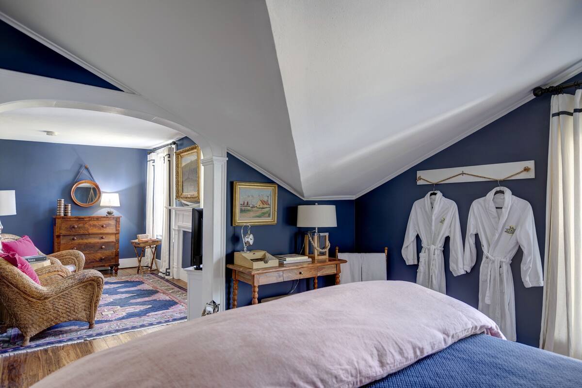 A bedroom with a large bed, a dresser, a chair, a table with a lamp, and two white robes hanging on the wall.