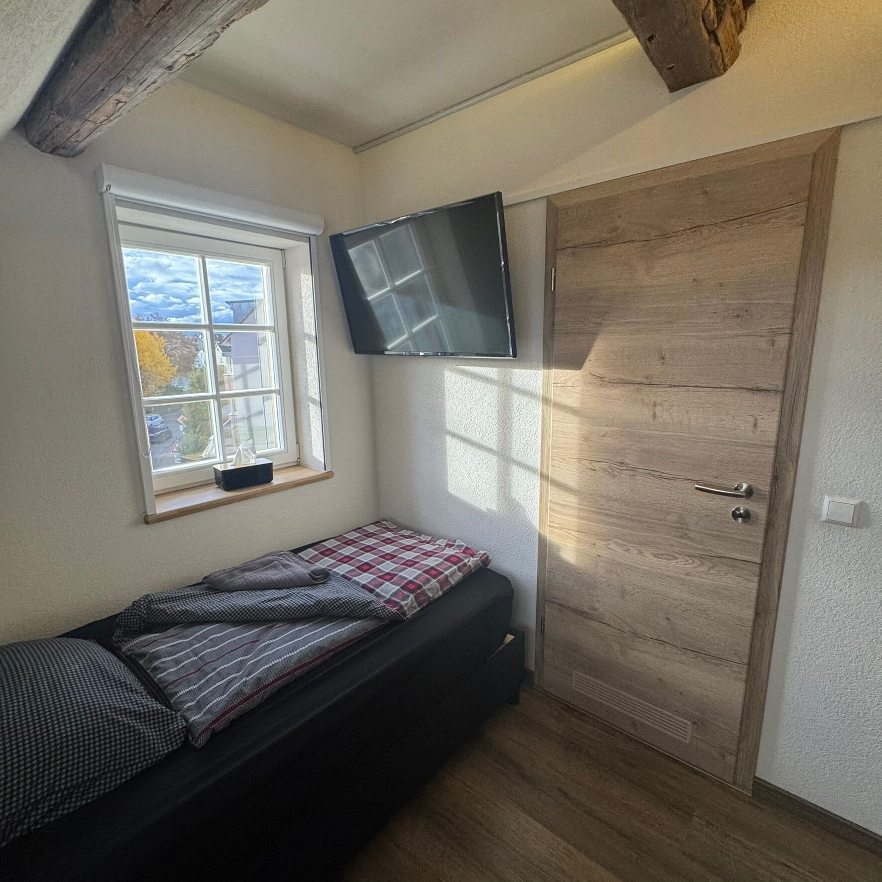 A small bedroom with a single bed, a window with a view, a wooden door, and a mounted TV.