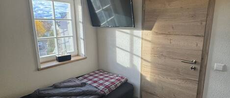 A small bedroom with a single bed, a window with a view, a wooden door, and a mounted TV.