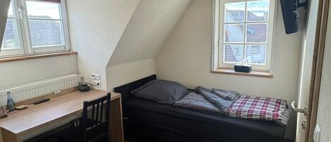 A small, triangular attic bedroom with a wooden desk, a bed, and a window overlooking a residential area.