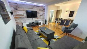 A modern living room with a stone wall, a flat-screen TV, a sofa set, a coffee table, and a dining area with chairs and a table. - Apartment Pallaton (Nasice)