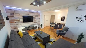 A modern living room with a stone wall, a flat-screen TV, a gray sofa with yellow cushions, and a dining area in the background. - Apartment Pallaton (Nasice)