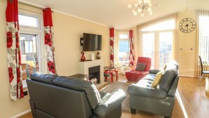 Een woonkamer met twee grijze banken, een rode fauteuil, een flatscreen televisie, een open haard en een groot raam met gordijnen.