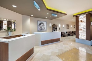 A hotel reception area with a reception desk, a large mirror, a potted plant, a framed artwork, and a seating area with chairs and tables.