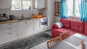 Een kleine keuken met witte kastjes, een spoelbak en een kookplaat. Er staat een rode bank en een eettafel met stoelen.