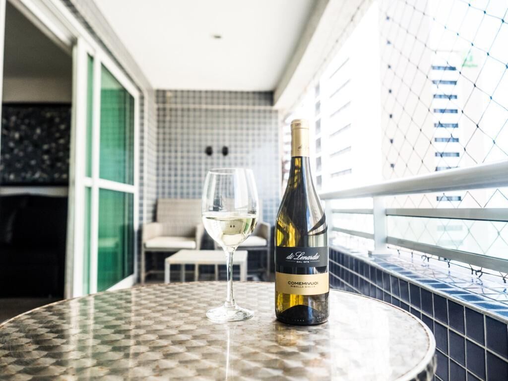 A bottle of wine and a glass on a round table with a checkered pattern, set in a room with a tiled wall and a balcony railing.