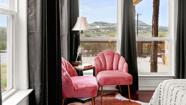 Une pièce avec deux fauteuils roses, une table avec une lampe et une vue sur un paysage vallonné, visible à travers de grandes fenêtres.