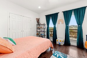 Quarto com cama, mesa de cabeceira, janela com cortinas, estante de livros e vista para o exterior.