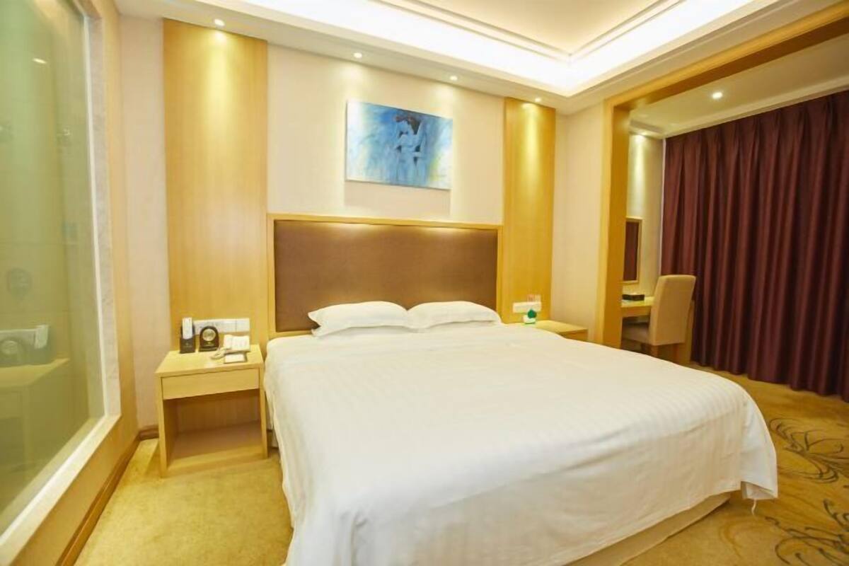 A hotel room with a large bed, a nightstand, a bedside table, a painting on the wall, and a window with curtains.