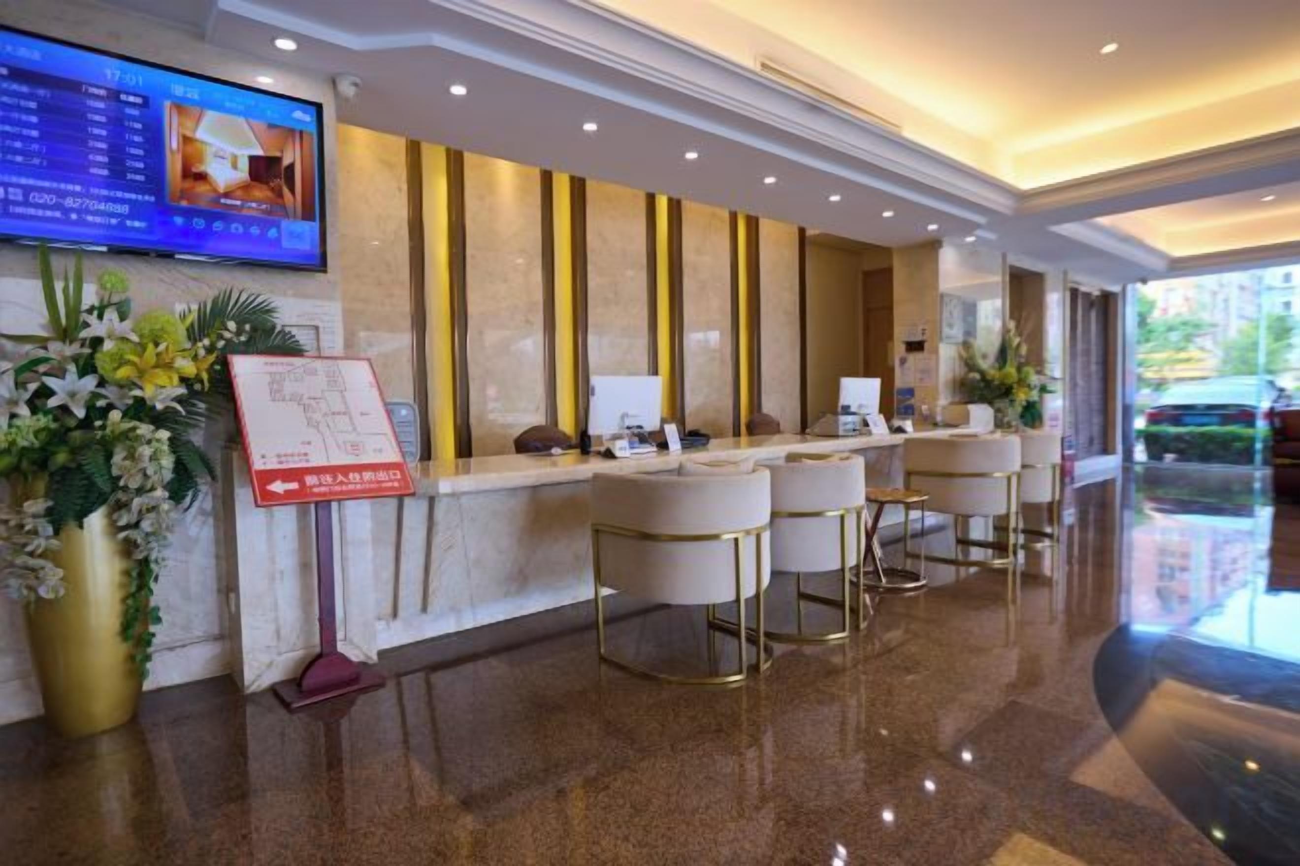 A hotel reception area with a digital screen, a reception desk, bar stools, and a plant.