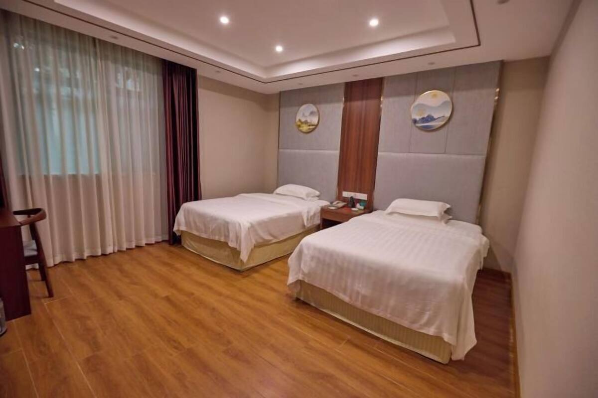 A hotel room with two single beds, a wooden floor, a large window with curtains, and a wall-mounted artwork.