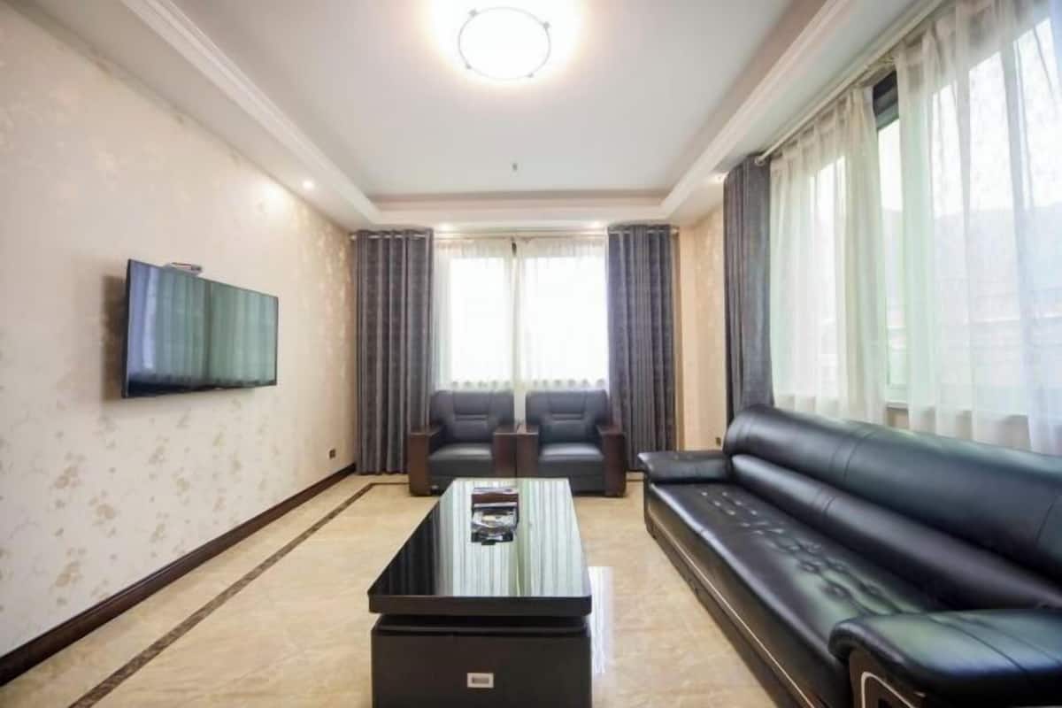 Una sala de estar con una televisión de pantalla plana montada en la pared, un sofá de cuero negro, dos sillones, una mesa de centro y un ventanal con cortinas.