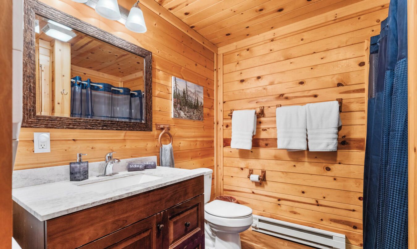 Un baño de cabaña de madera con tocador de madera, lavabo, espejo, inodoro y toallas colgadas en la pared.