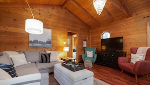 Ein gemütliches Blockhaus-Interieur mit einer Couch, einem Hocker, Sesseln, einem Fernseher und einer Chandelier.