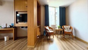 A room with a wooden desk, a television, two chairs, a small table, and a window with curtains.