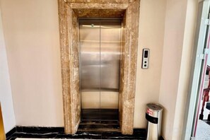 Een lift met een metalen afwerking, een bedieningspaneel aan de muur en een prullenbak aan de rechterkant.