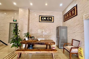 A hotel lobby with a reception desk, a wooden bench, a chair, a potted plant, and a refrigerator.