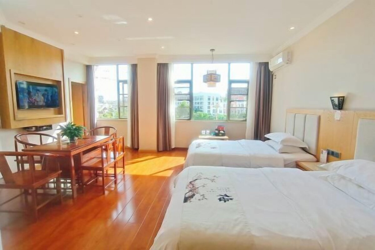Ein Hotelzimmer mit zwei Betten, einem Esstisch mit Stühlen, einem Fernseher und einem großen Fenster mit Blick auf die Stadt.