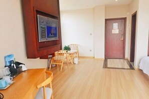 Een hotelkamer met een houten vloer, een eettafel met stoelen, een aan de muur bevestigde televisie en een deur met een deurmat.