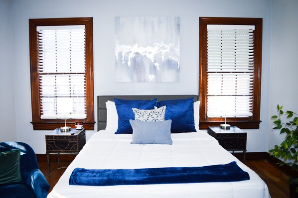 A bedroom with a large bed, two nightstands, a wall-mounted artwork, and a window with blinds.