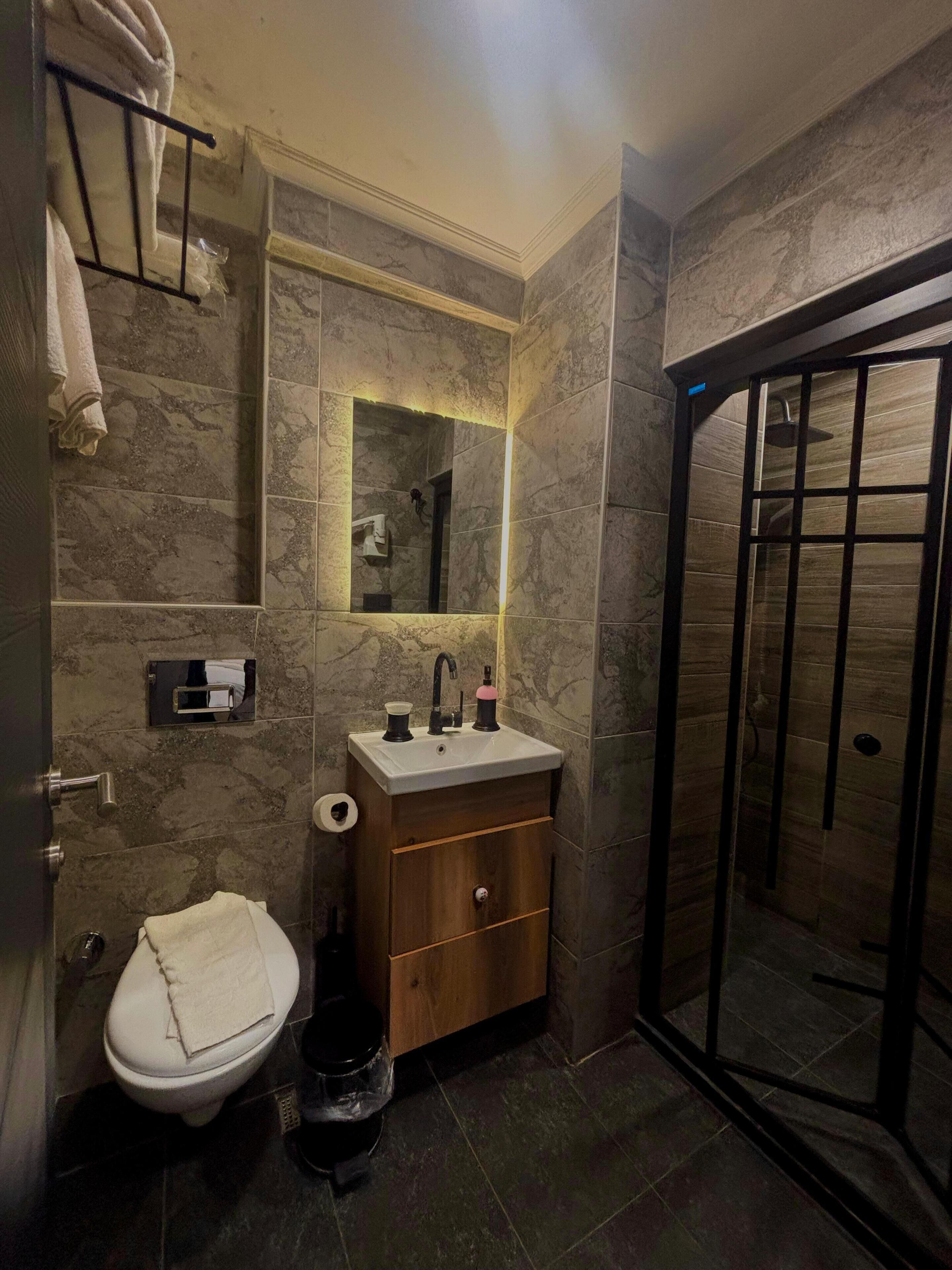 Une salle de bain avec un mur en marbre, un meuble-lavabo en bois, un lavabo blanc, des toilettes et une cabine de douche en verre.