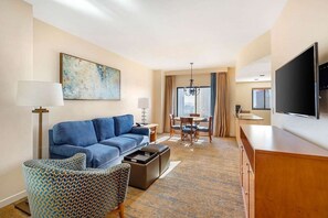 Une chambre d’hôtel moderne avec un canapé bleu, un fauteuil à motifs, un coin repas comprenant une table et des chaises, et un téléviseur fixé au mur.