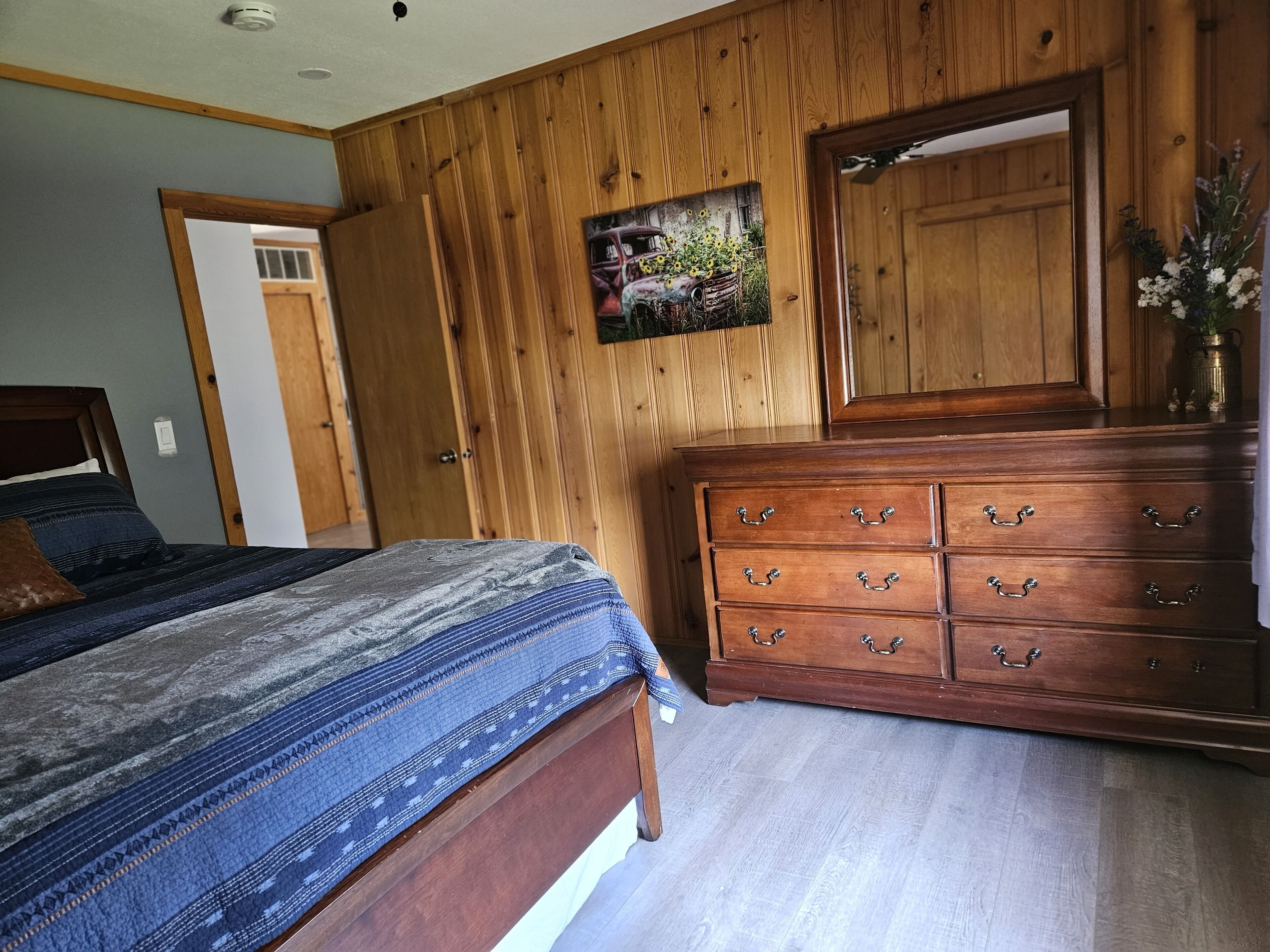 A bedroom with a wooden headboard, a bed with a patterned blanket, a wooden dresser with drawers, and a painting on the wall.