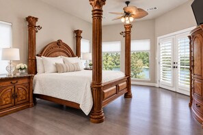 A four-poster bed with a wooden headboard and footboard, a nightstand with a lamp, and a view of greenery through the windows.
