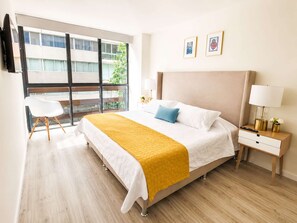 A hotel room with a large bed, a bedside table with a lamp, a chair, and a window with a view of buildings.