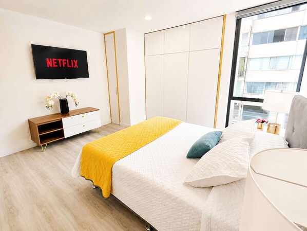 A modern bedroom with a bed, a TV mounted on the wall, a wardrobe, and a window with a view of buildings.