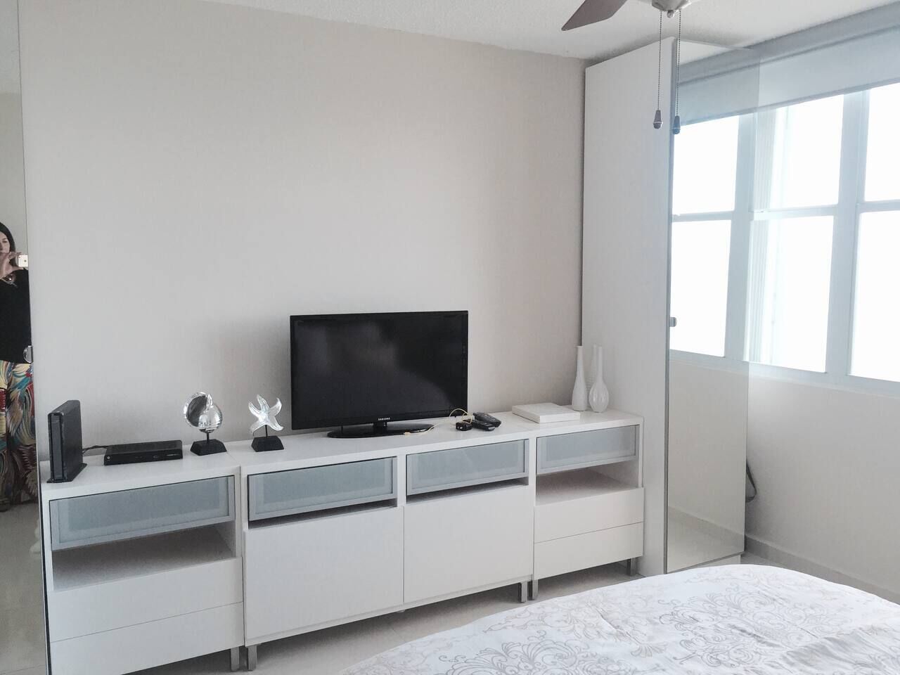 A bedroom with a bed, a TV on a white dresser, a window with blinds, and a ceiling fan.