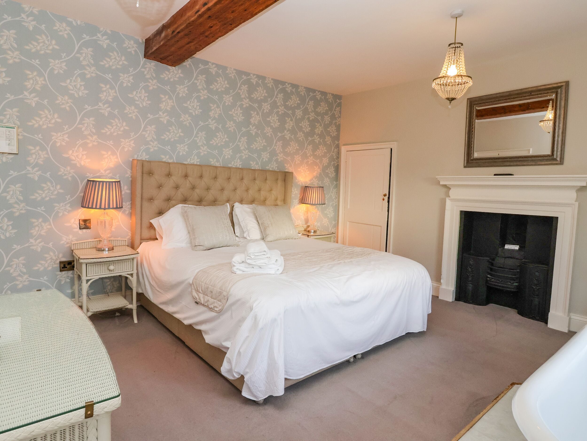 A neatly arranged bedroom with a large bed, two bedside lamps, a mirror above the fireplace, and a patterned wallpaper.