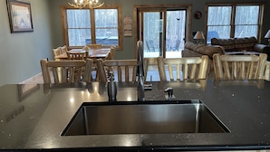A kitchen with a central island, wooden chairs, a dining table, and a view of the outdoors through large windows.