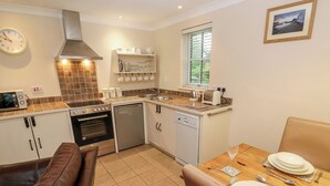A compact kitchen with a dining area, equipped with a stove, oven, dishwasher, and a small window. - Tregonning Lodge (Helston)