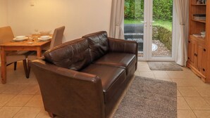 A living room with a brown leather sofa, a dining table set with plates and glasses, a wooden cabinet, and a sliding glass door leading to a garden. - Tregonning Lodge (Helston)
