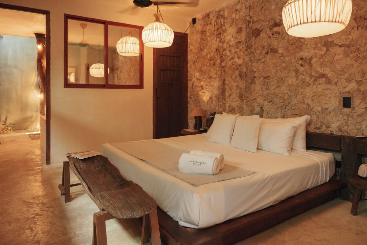 Une chambre avec un mur en pierre, un cadre de lit en bois, un banc et deux luminaires suspendus.