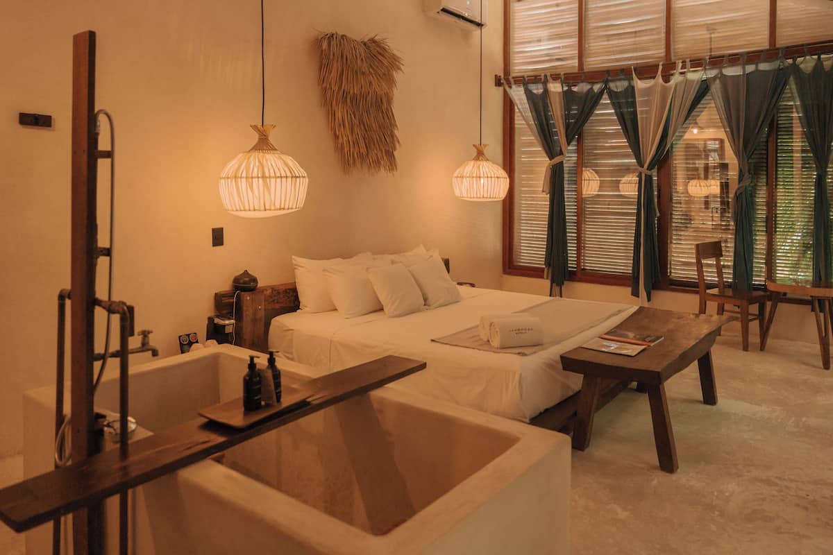 Une chambre d’hôtel avec un grand lit, une table de chevet en bois, une petite table en bois, une baignoire et une fenêtre avec des rideaux.