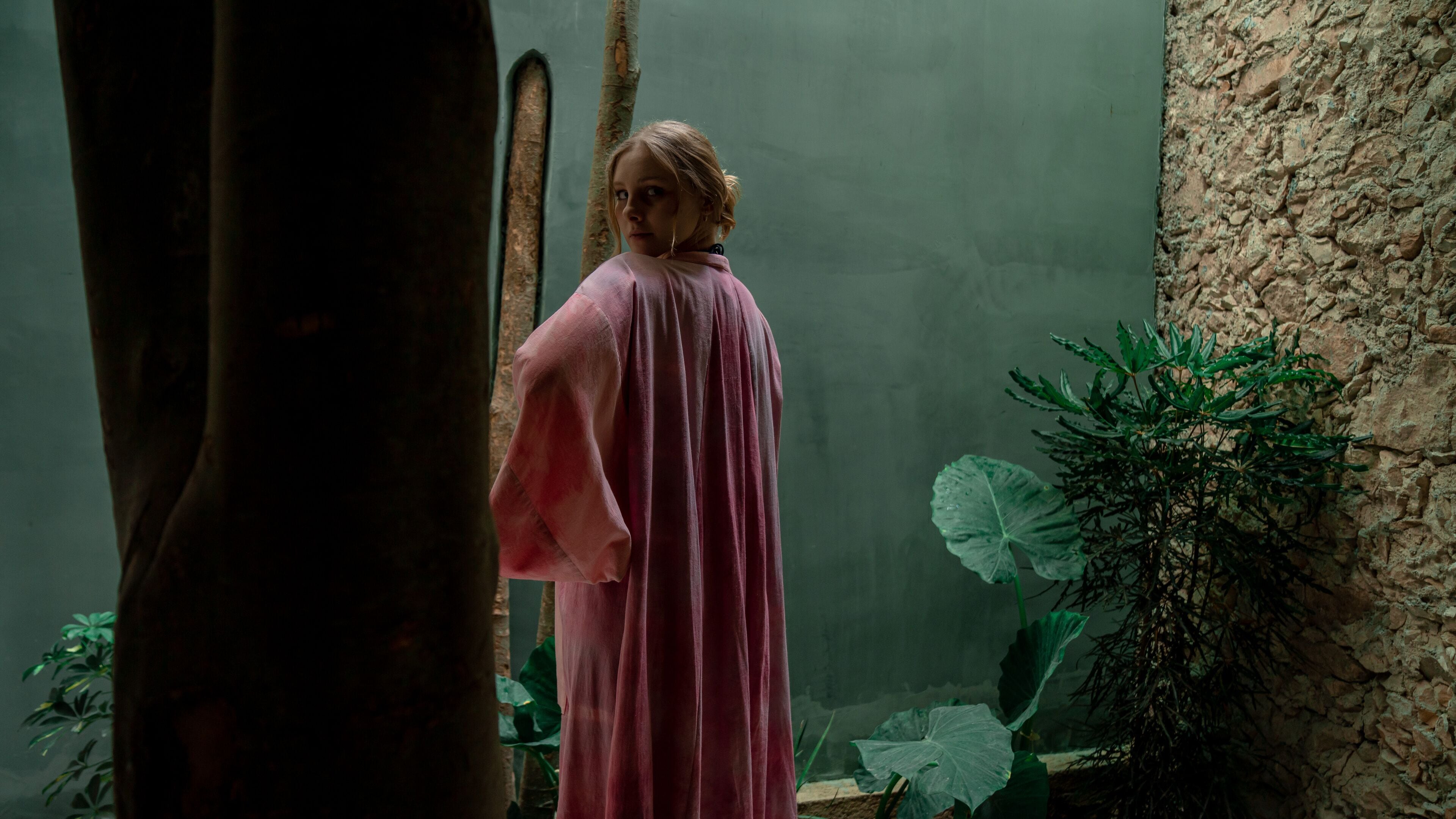 A person in a pink robe standing in a room with a stone wall and large green leaves.