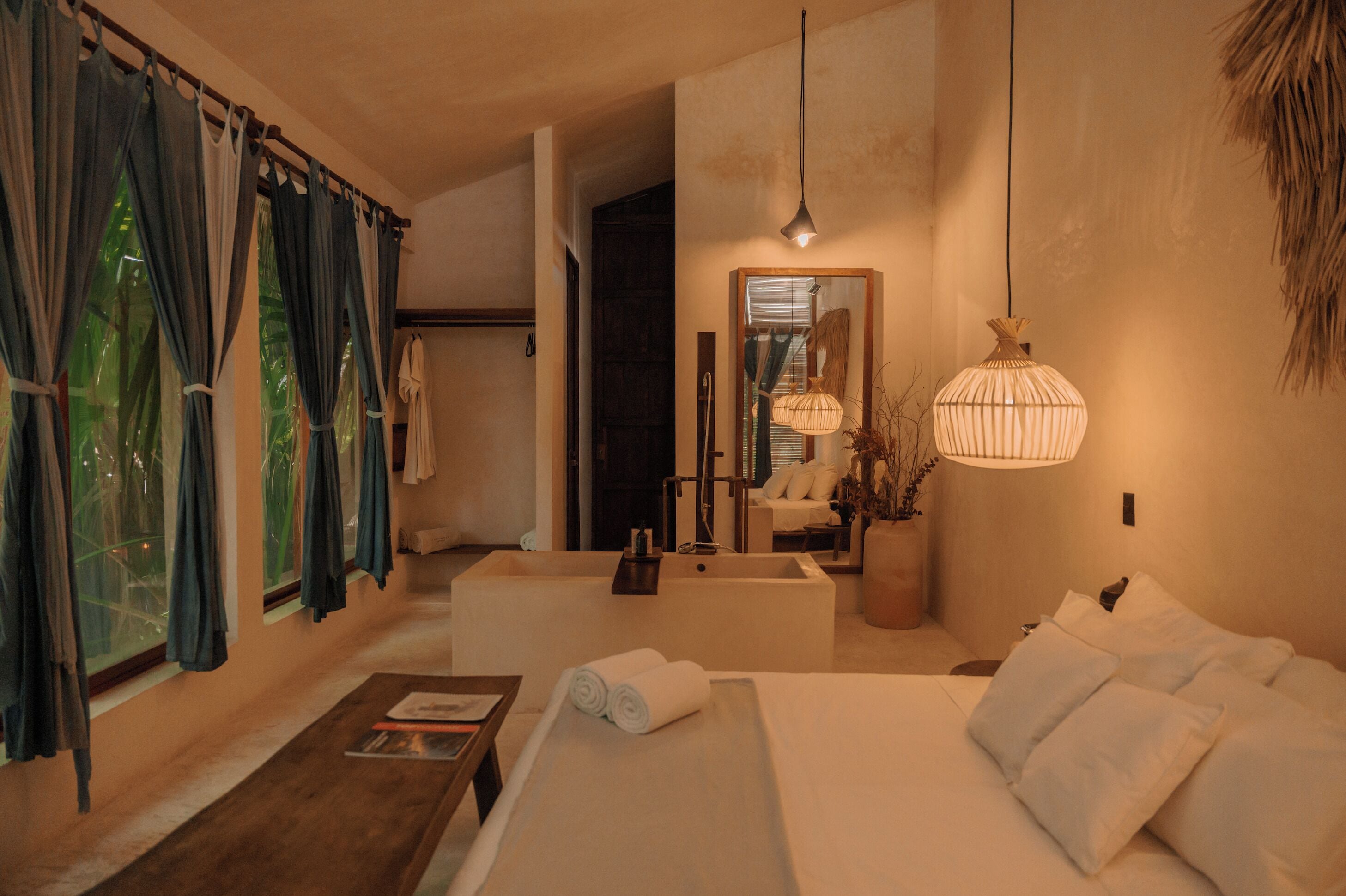 A modern hotel room with a large bed, a sitting area with a table, a bathtub, and a large window with curtains.