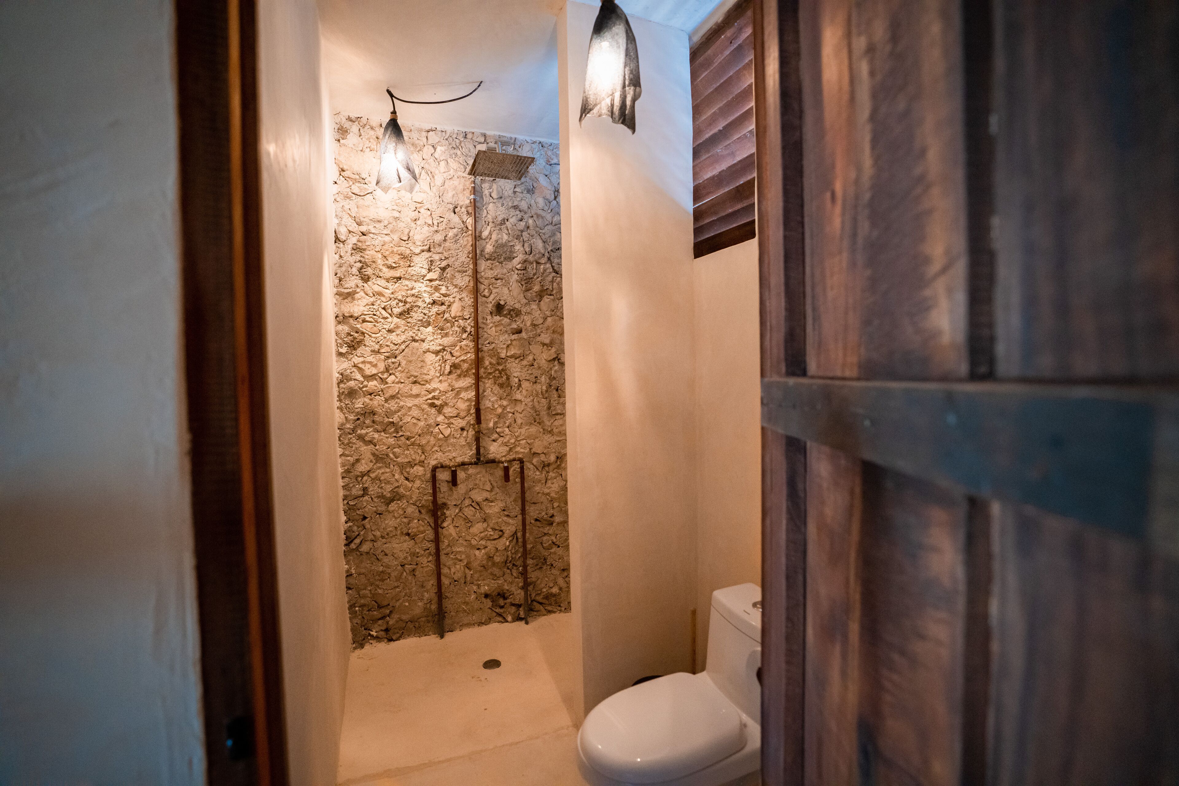 A bathroom with a stone shower wall, a wooden door, and a white toilet.