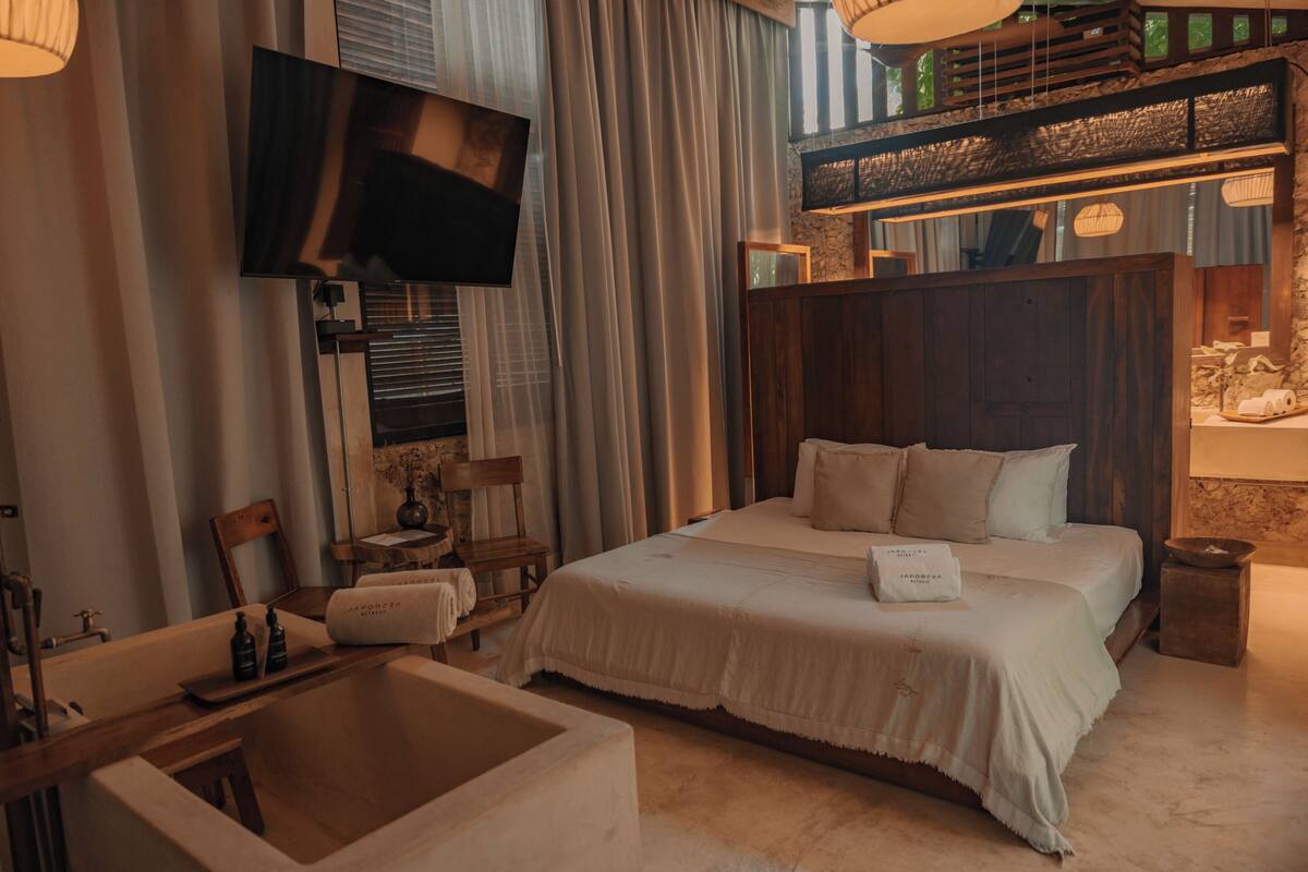 Une chambre d’hôtel avec un grand lit, un téléviseur fixé au mur, une tête de lit en bois, une petite table avec une bouteille et une fenêtre avec des rideaux.