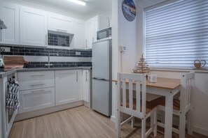 A modern kitchen with white cabinets, a built-in microwave, and a refrigerator. There is a small dining table with a chair and a decorative wall clock. - Castle Cove - 1 Bedroom Apartment - Tenby (Tenby)