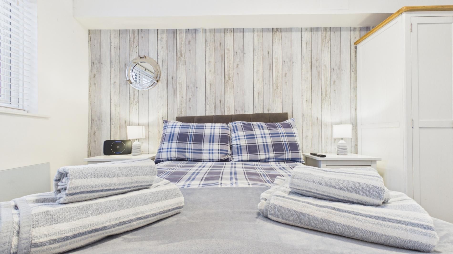 A bedroom with a wooden wall panel, a round mirror, a bed with plaid bedding, two bedside tables with lamps, and a door.