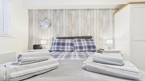 A bedroom with a wooden wall panel, a round mirror, a bed with plaid bedding, two bedside tables with lamps, and a door. - Castle Cove - 1 Bedroom Apartment - Tenby (Tenby)