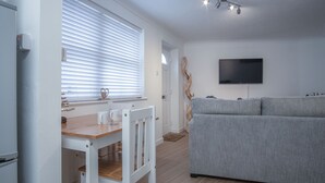 Un salon moderne avec un canapé gris, une table à manger en bois avec des chaises, un téléviseur à écran plat fixé au mur et une fenêtre avec des stores.